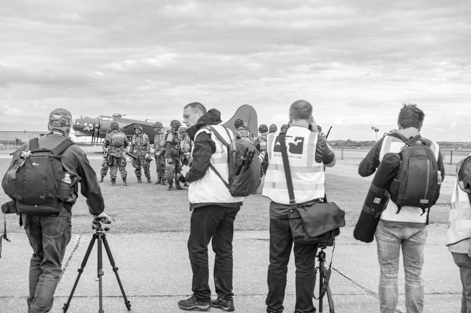 Media journalists with cameras and equipment wait in a black and white photo at an outdoor event location.