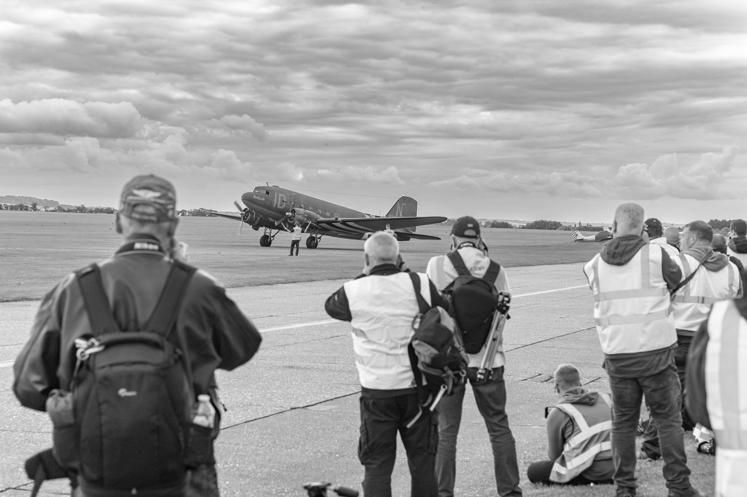 Spectators watching vintage aircraft on airfield tarmac with Douglas DC-3 transport plane in background.
