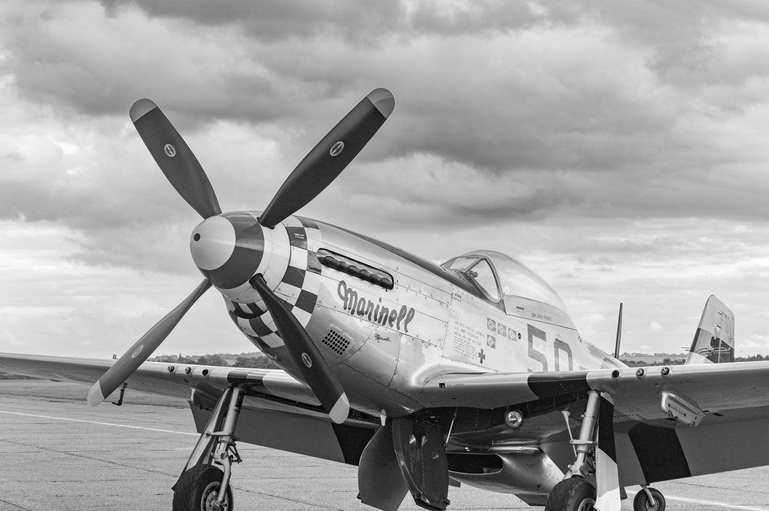 Black and white photo of a P-51 Mustang fighter aircraft with prominent propeller on display against cloudy sky.