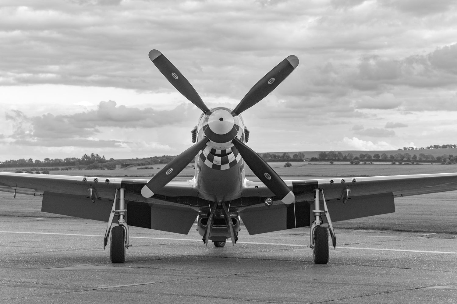 Vintage P-51 Mustang fighter aircraft with distinctive propeller and nose on an airfield under cloudy skies.