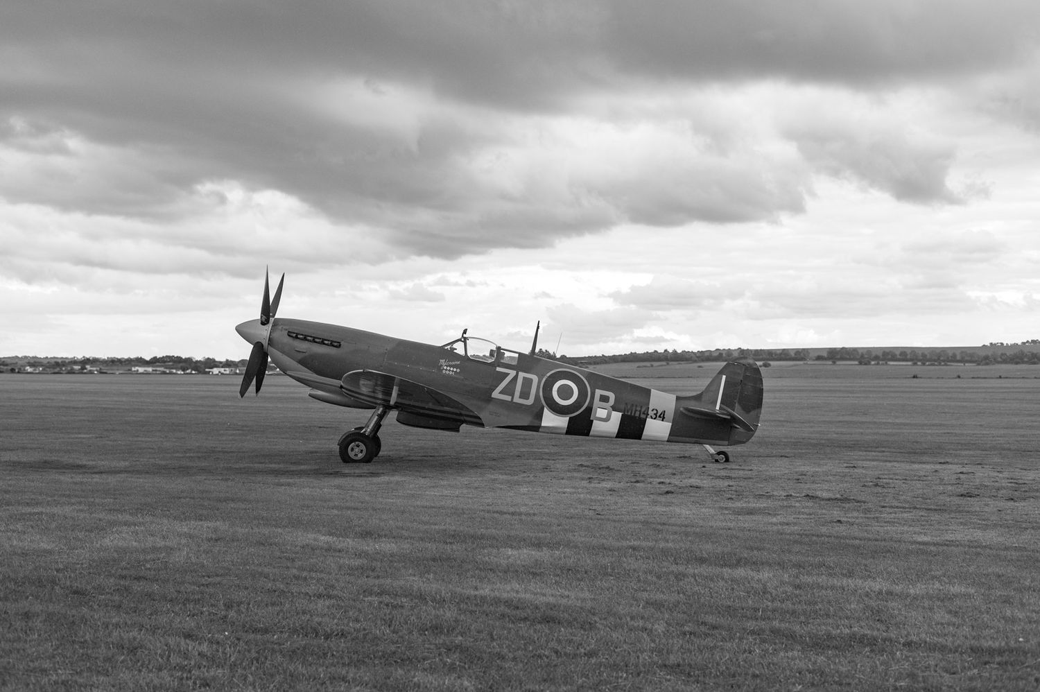 A vintage World War II Spitfire fighter plane taxis across a grassy airfield under dramatic cloudy skies.