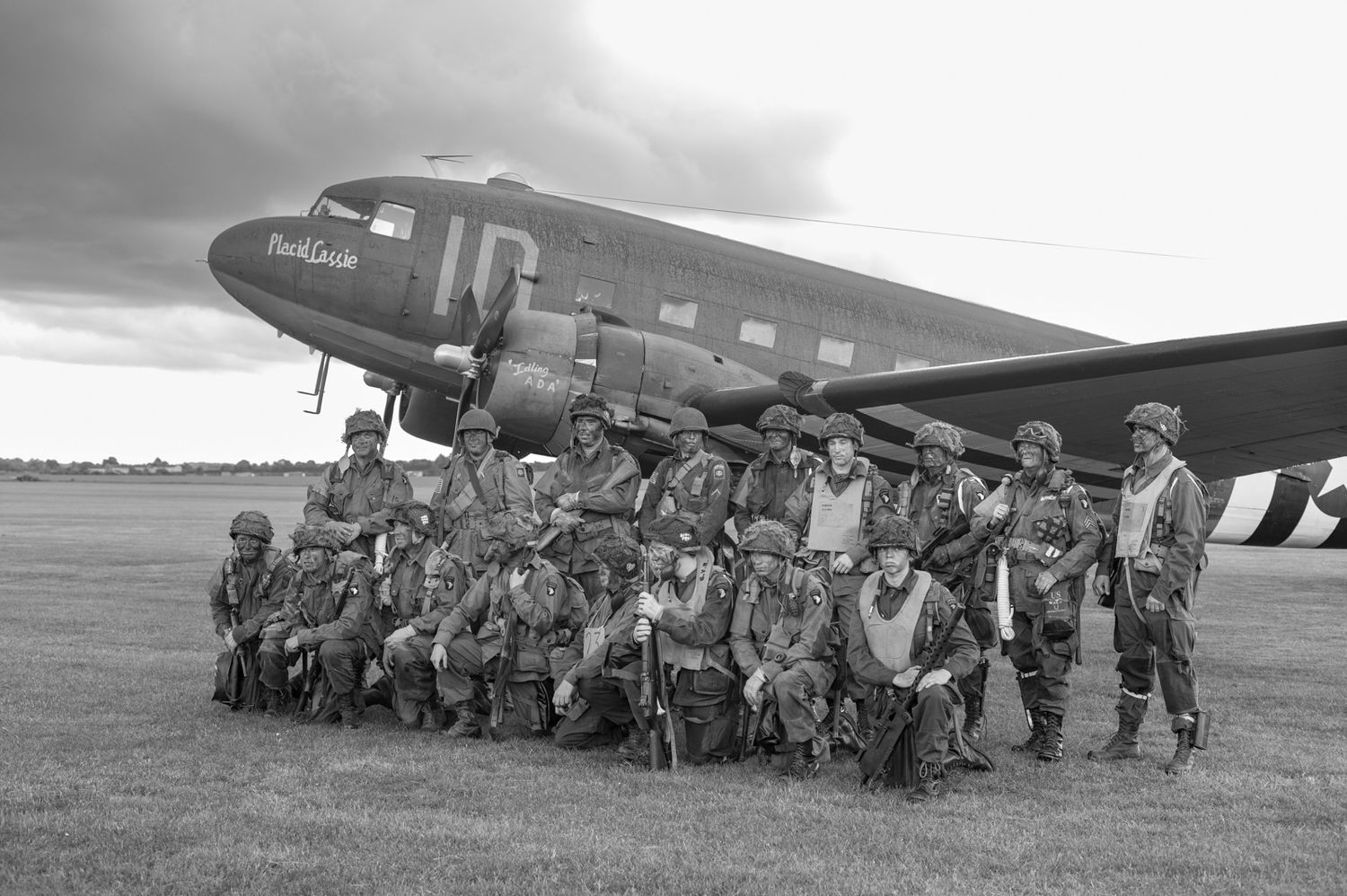 Military personnel gathered in formation beside vintage Douglas C-47 transport aircraft on grassy airfield.