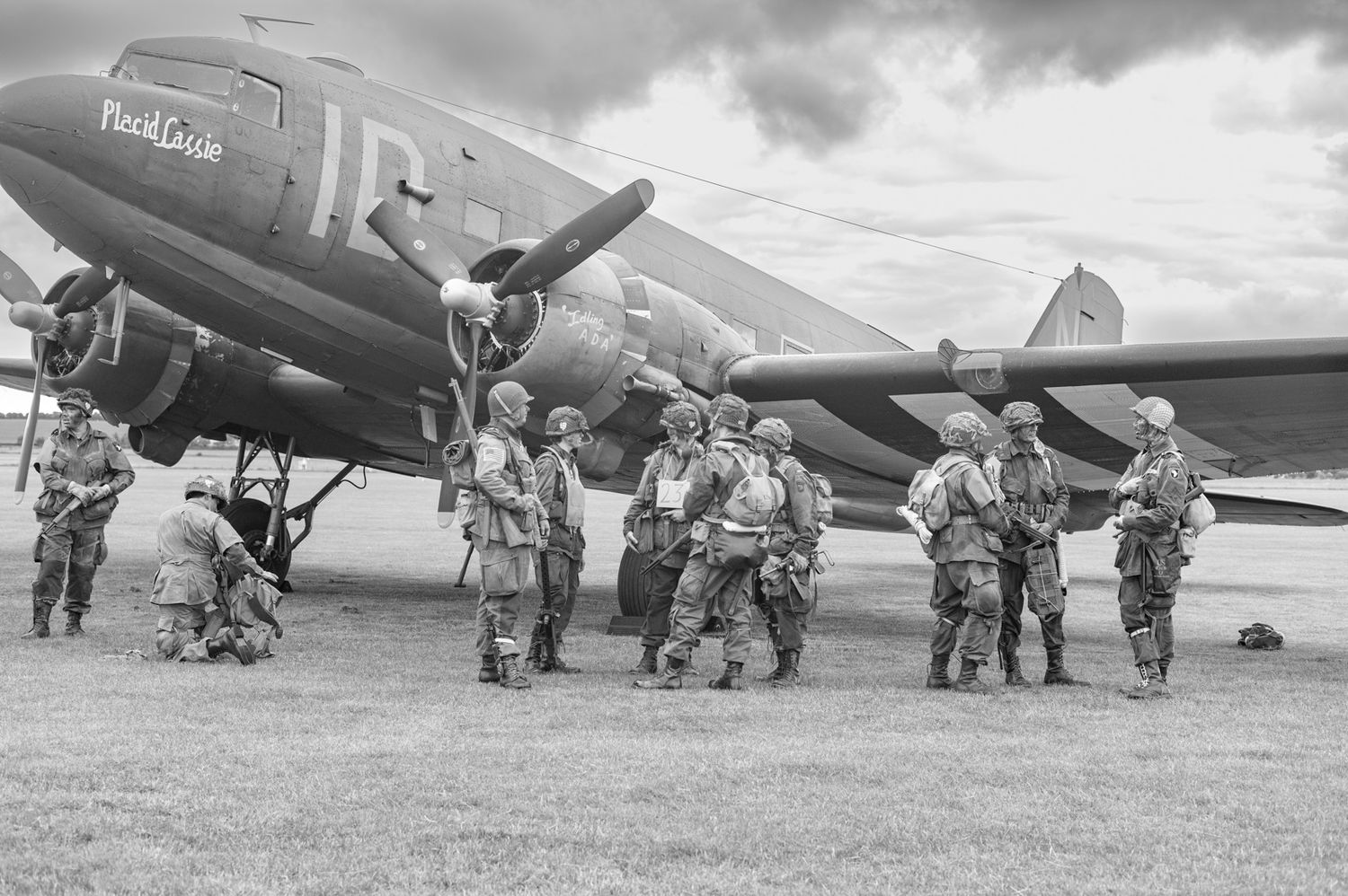 Military personnel stand alongside a historic twin-engine transport aircraft on an airfield in black and white.