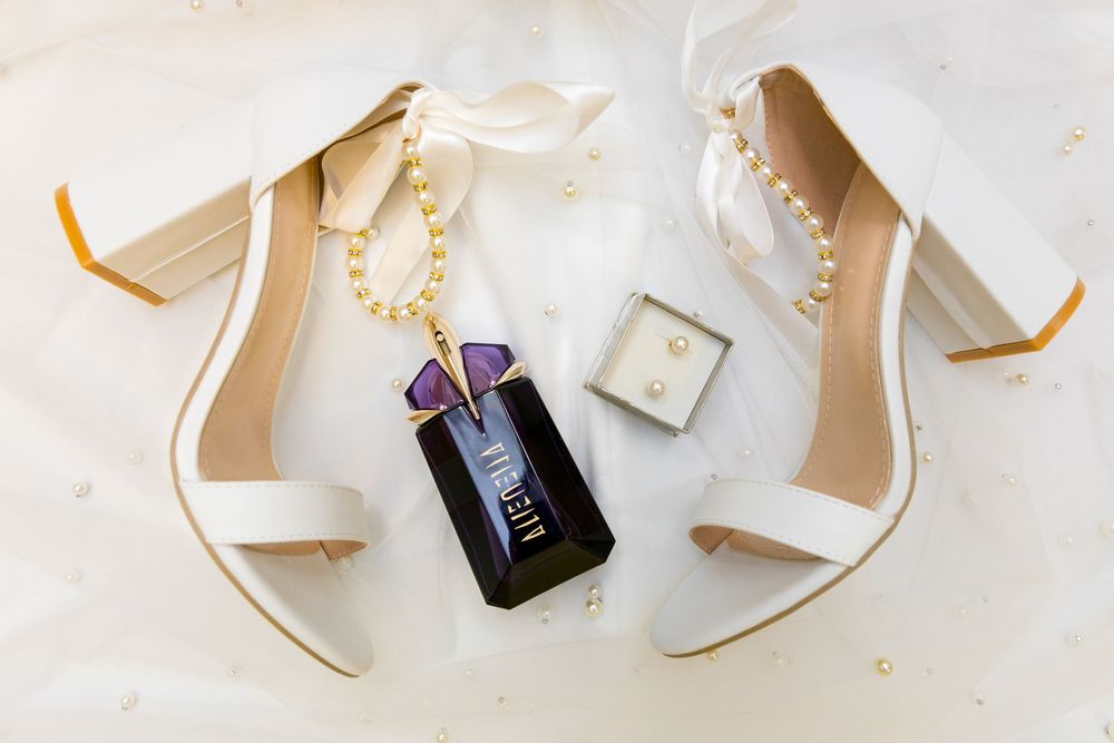 Bridal accessories including white heels, pearl jewellery and Alien perfume on soft fabric background.