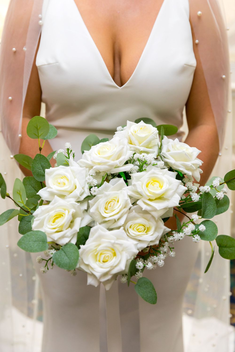 Bride in a white gown holding a bouquet of ivory roses and greenery with delicate pearl veil.
