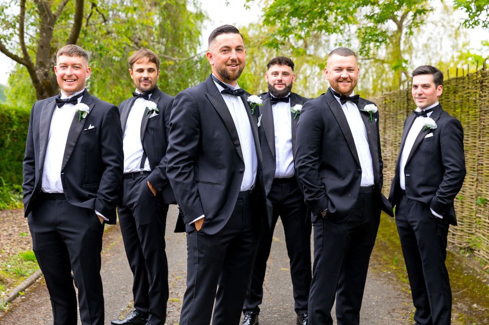 Groom and groomsmen in black tuxedos posing confidently outdoors before the ceremony.