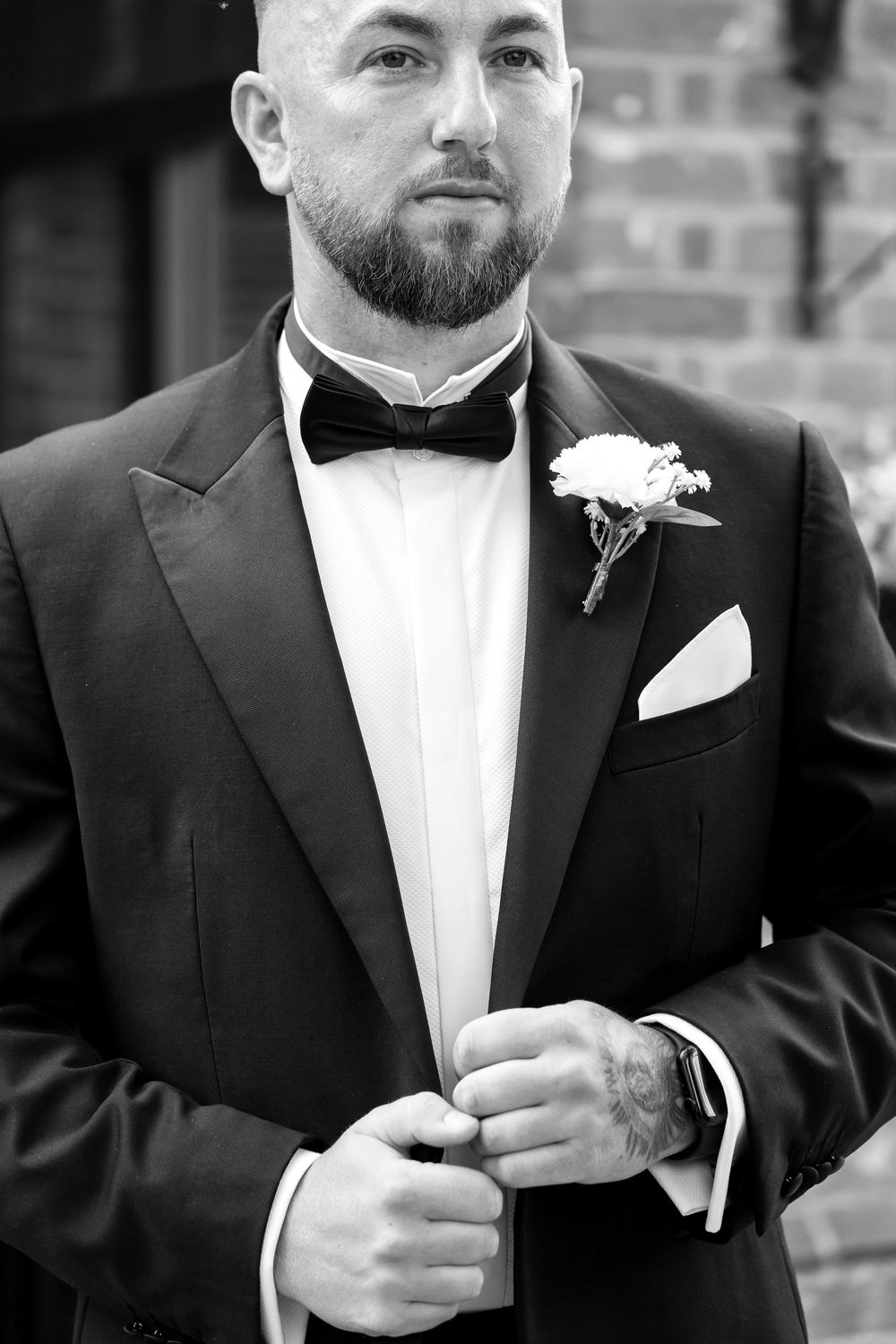 Black and white portrait of groom adjusting suit, wearing a bow tie and boutonnière.