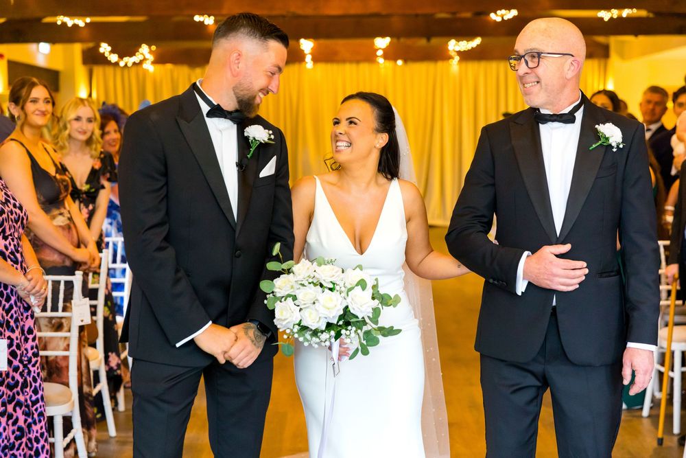 Bride walking down the aisle with father, smiling at groom, surrounded by happy guests.