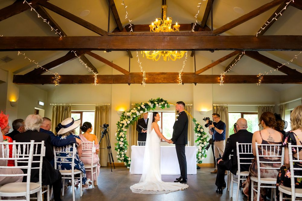 Wedding ceremony in rustic barn venue with floral arch and fairy lights as bride and groom exchange vows.