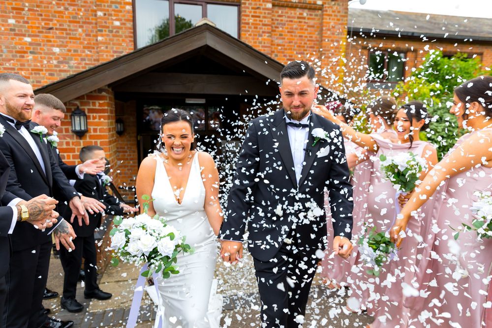 Bride and groom walk through confetti tunnel laughing as guests celebrate outside the venue.