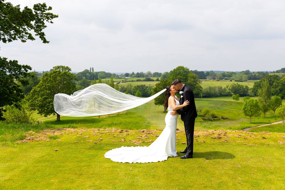 Bride and groom kiss on a hilltop as her veil flows in the wind with countryside views in the background.