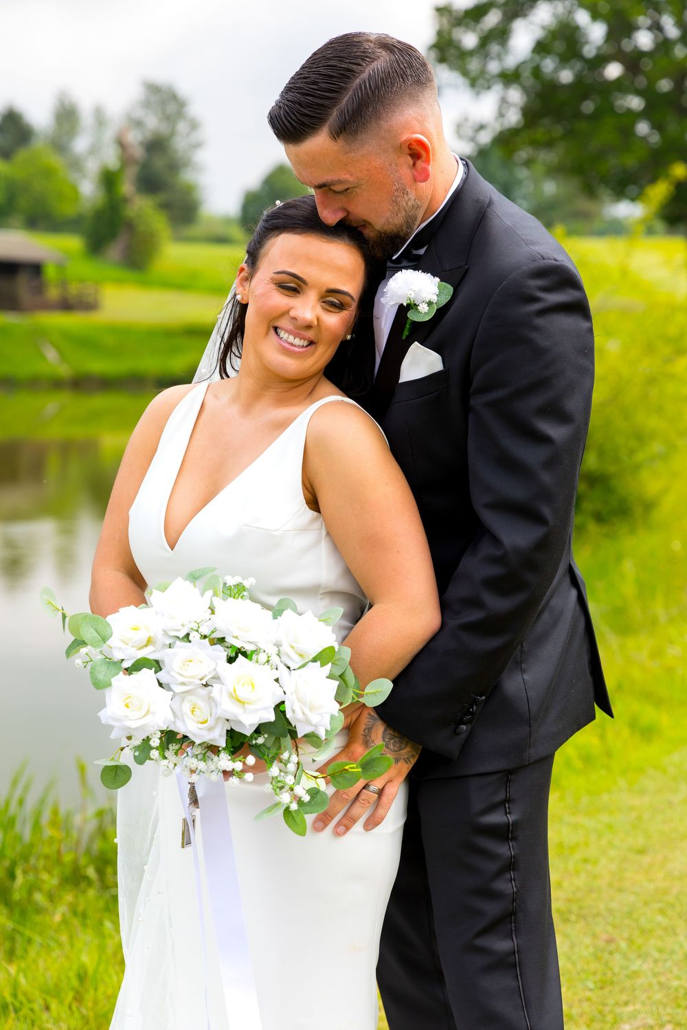 Bride smiles holding bouquet as groom kisses her head beside a pond on the golf course.