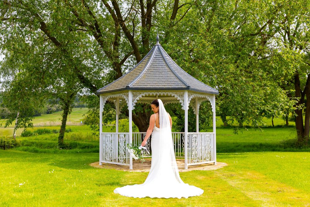 Bride stands with her back to the camera at a white garden feature surrounded by lush greenery.