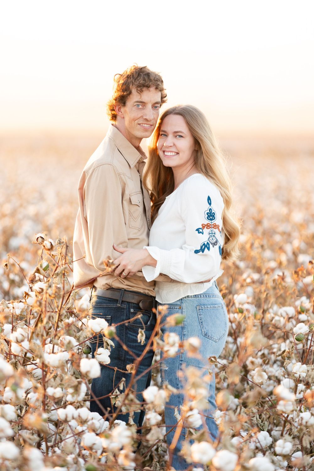 Couple embracing in a blooming cotton field during sunset with warm golden light surrounding them.