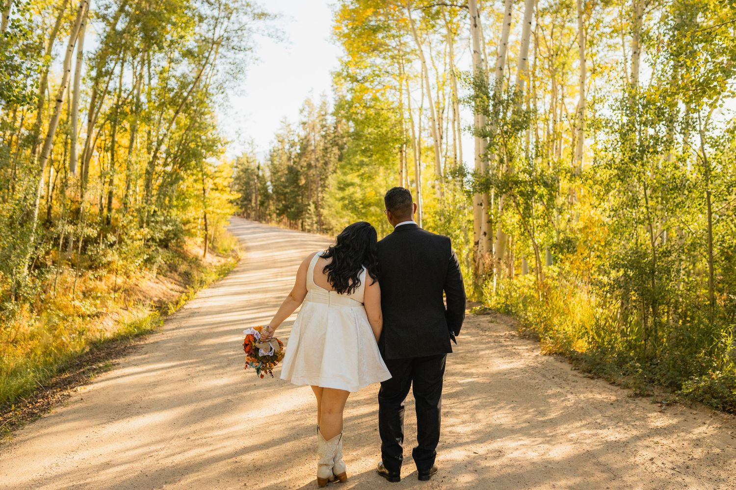 A couple walks down a sunlit dirt path surrounded by golden aspen trees on an autumn day.