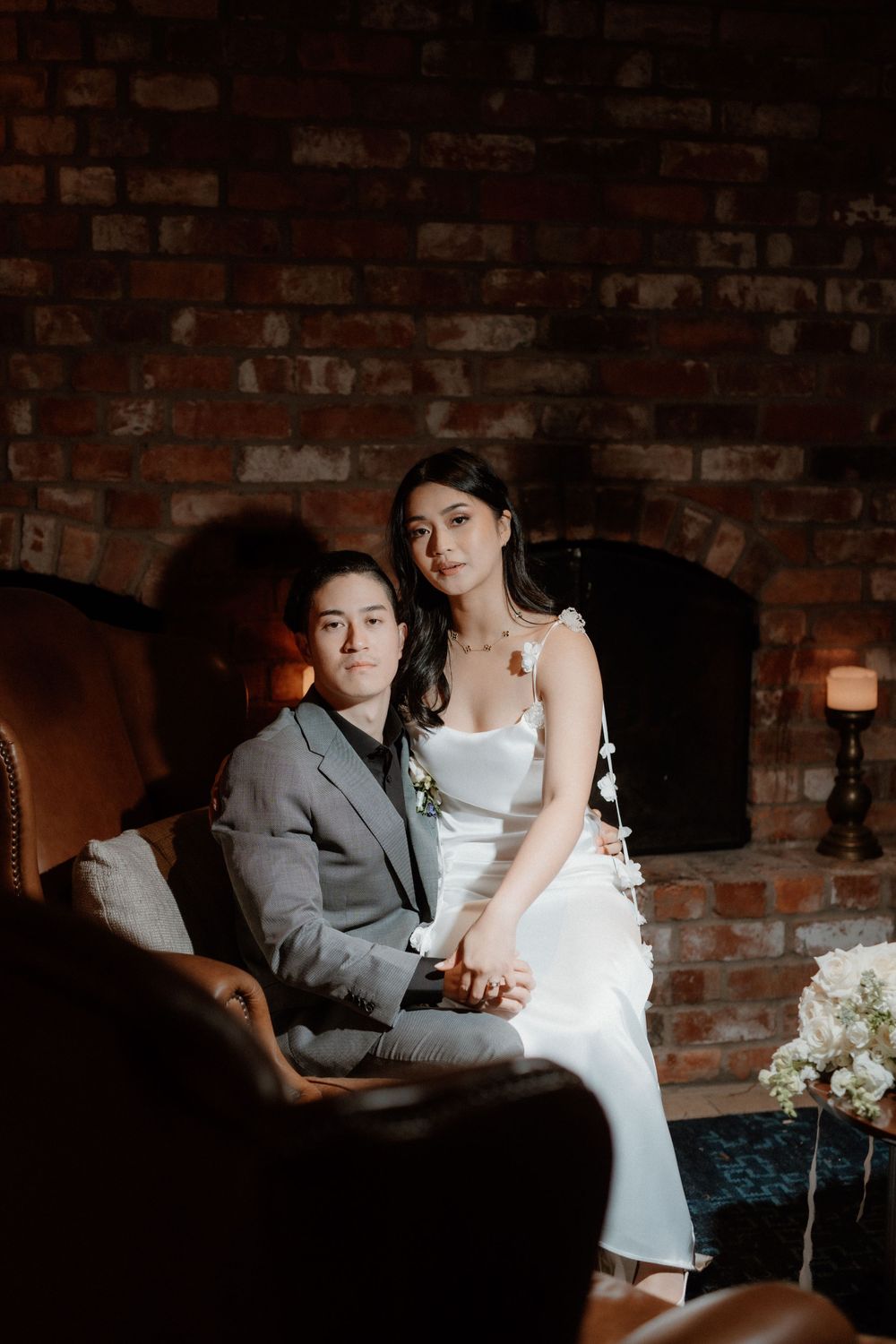 A couple in elegant wedding attire pose together on a vintage leather chair against a rustic brick wall with moody lighting.