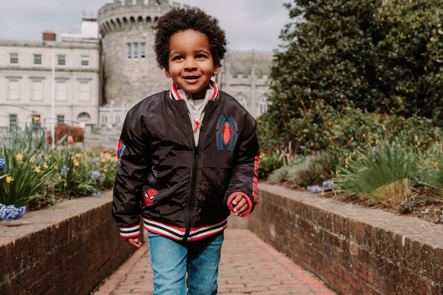 A child wearing a black varsity jacket and jeans stands smiling in front of a castle-like building with gardens.