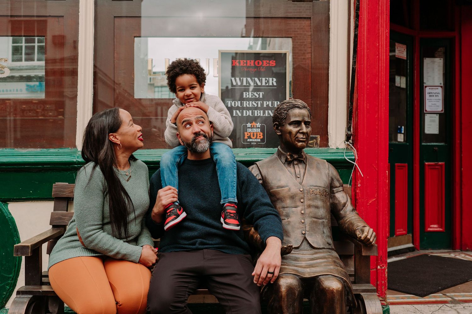 A family sits together on a bench next to a bronze statue in front of a storefront with red trim.