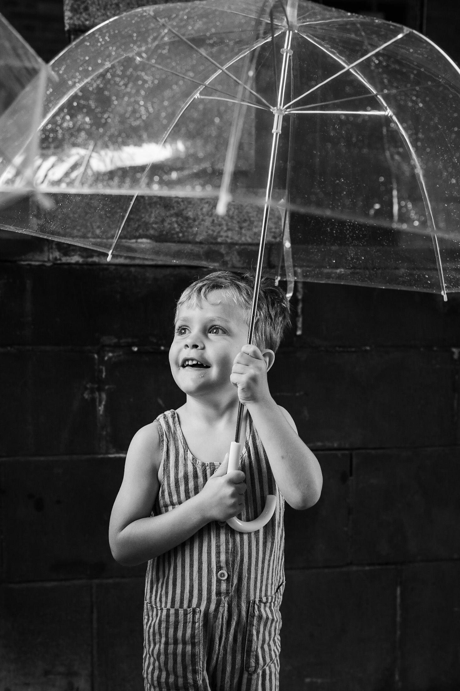 A vintage black and white photograph of a small child in striped overalls holding up a clear umbrella while looking up.