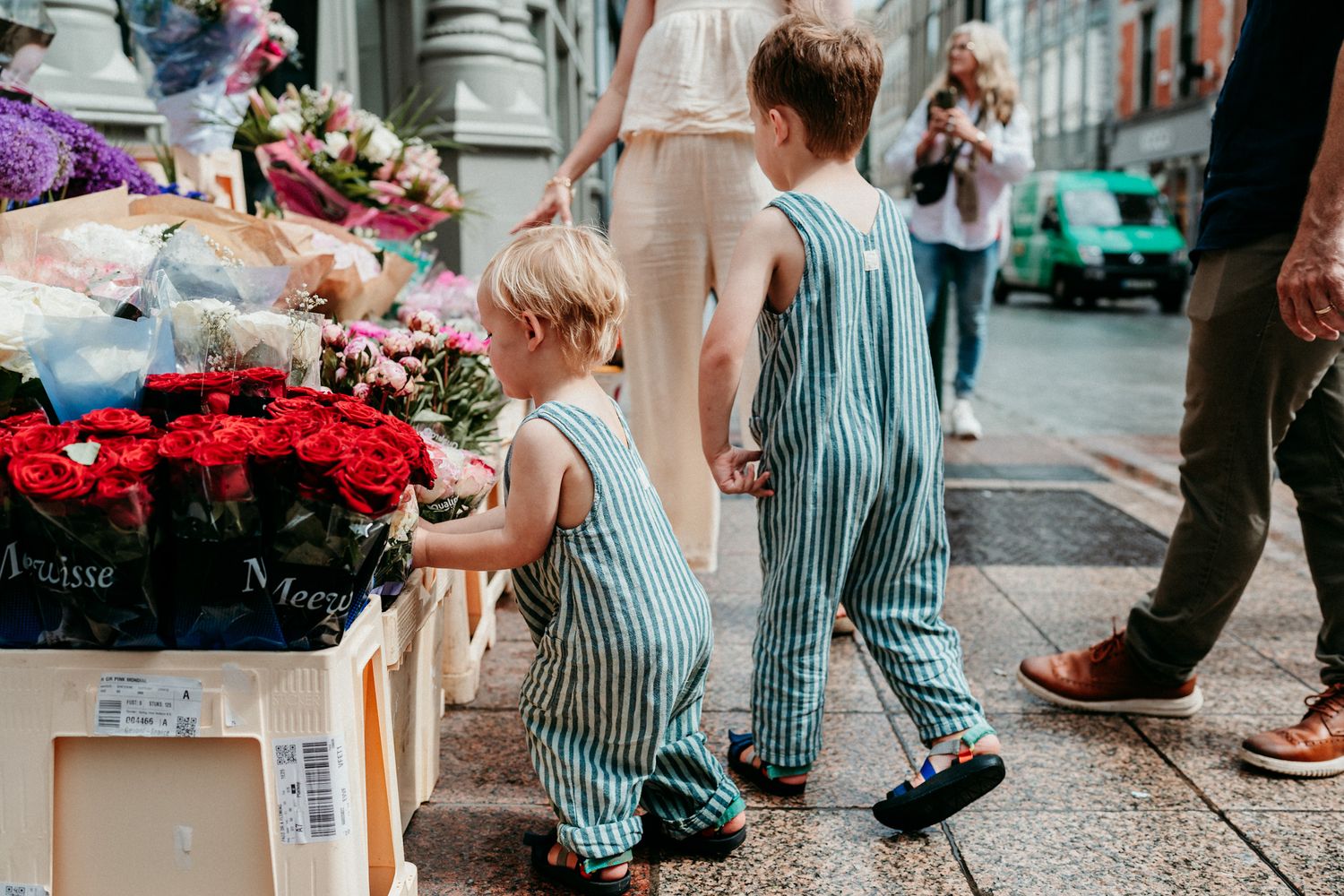 Children in striped overalls examine colorful flowers at an outdoor market stall.