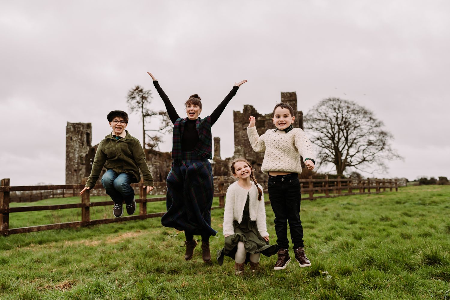Four children jump and play in a green countryside field with wooden fences and trees in the background.