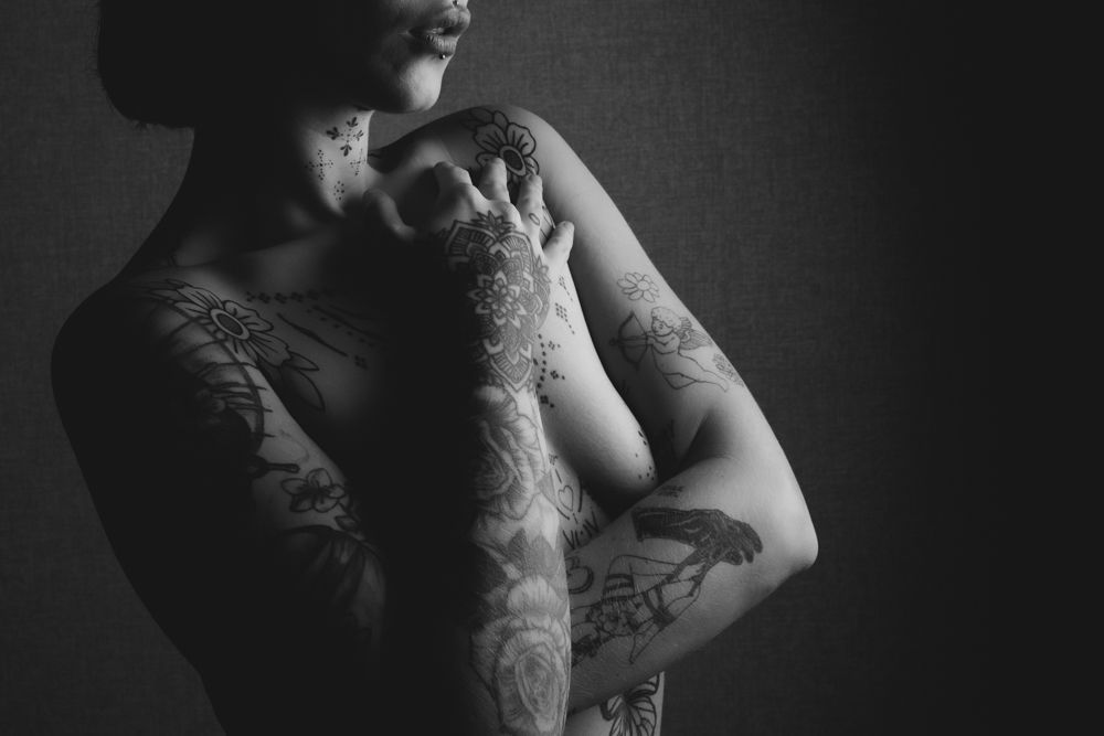 Dramatic black and white photograph showing tattooed arms crossed against torso in moody lighting.