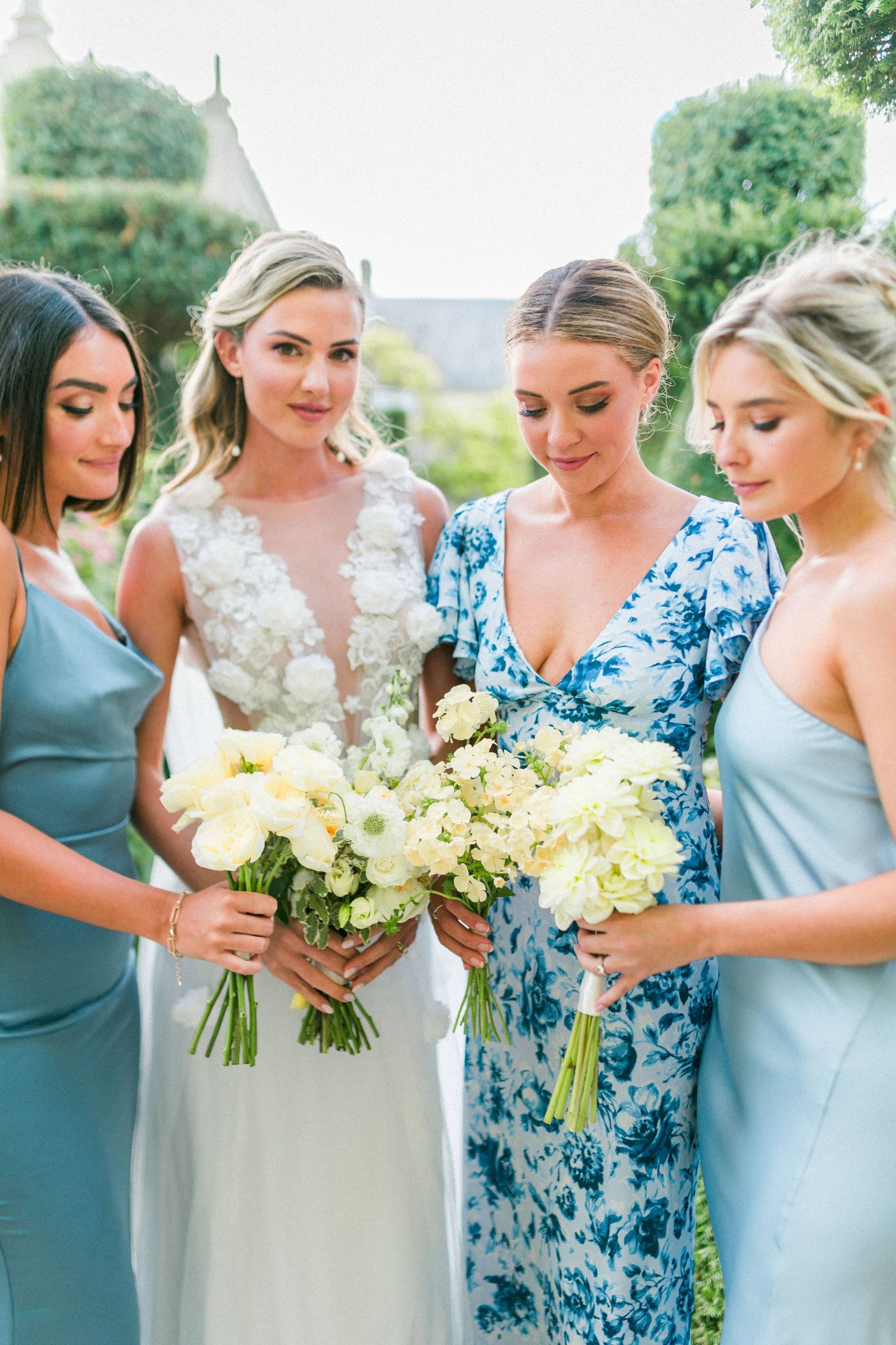 A bridal party in pastel blue and floral dresses admire white and cream bouquets together at Euridge Wiltshire - Hayley Elizabeth Photograph
