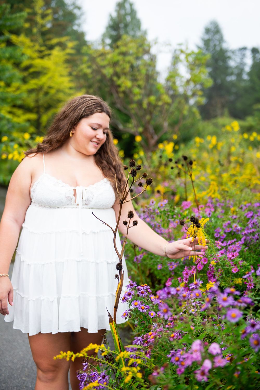 Woman in white sundress poses gracefully in a garden filled with vibrant yellow and purple wildflowers on a sunny day.