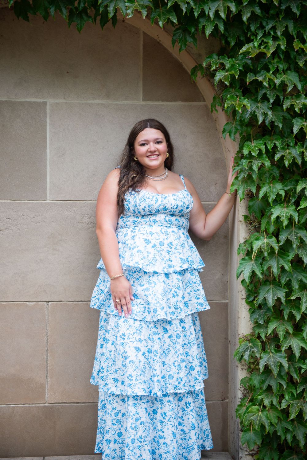 A person in a tiered blue and white floral summer dress stands in an archway framed by climbing ivy.