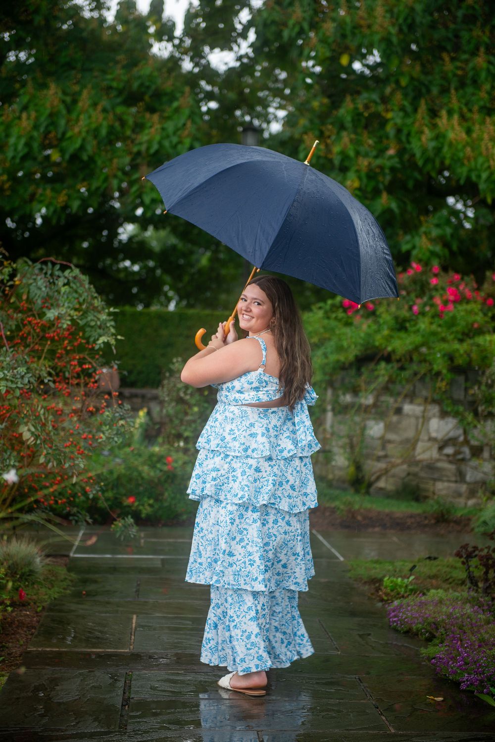 A person in a blue floral tiered dress poses with a navy umbrella on a wet stone path surrounded by garden foliage.
