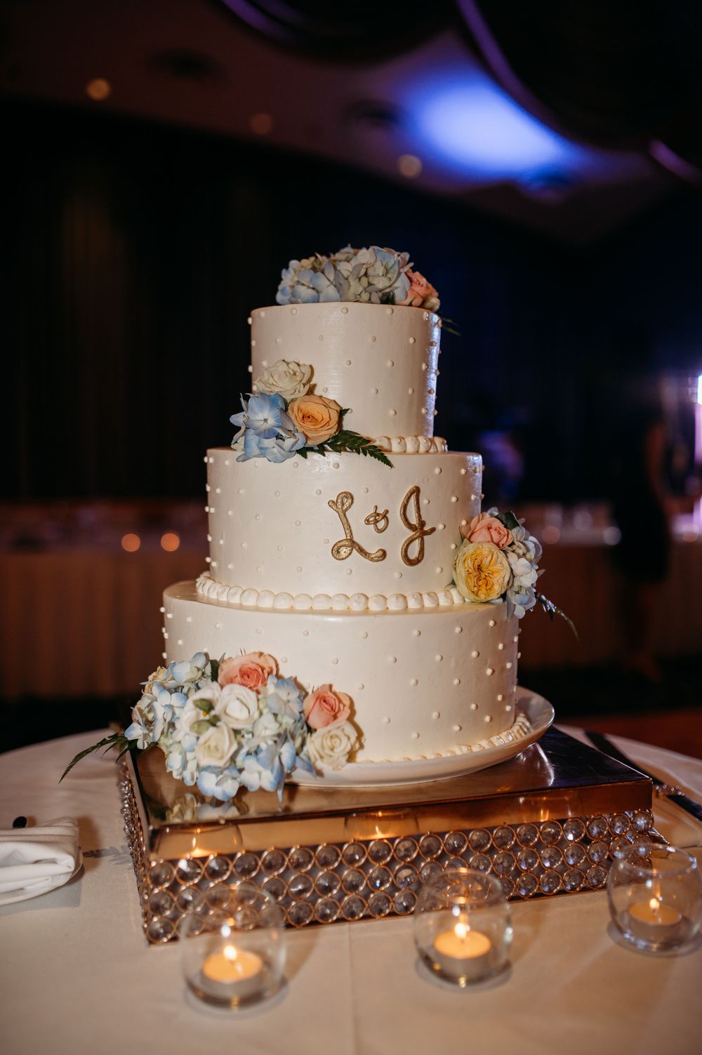 Three-tiered white wedding cake with blue and peach flowers on crystal stand.