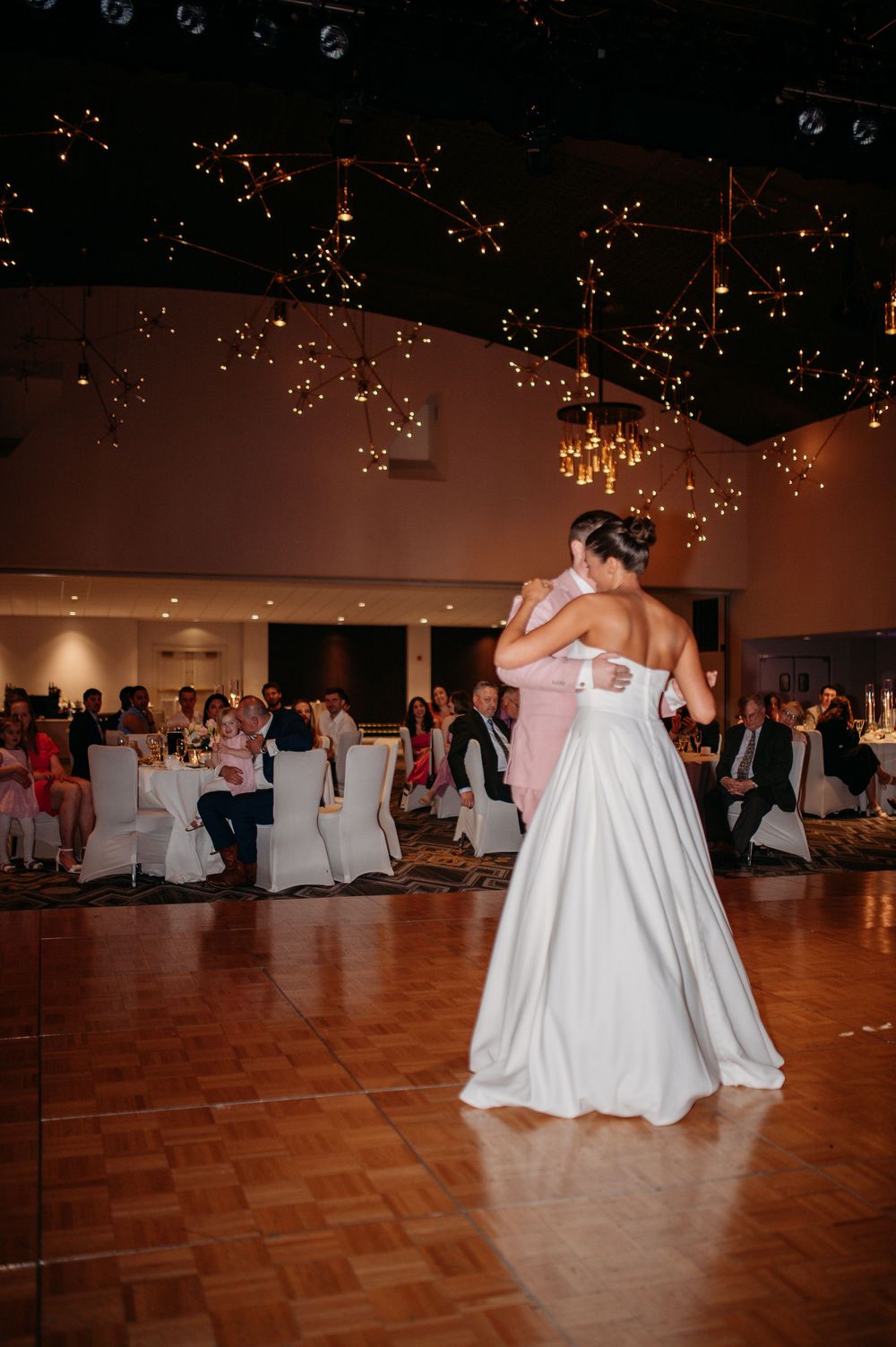 Couple shares intimate first dance on wooden floor under string lights at wedding reception.
