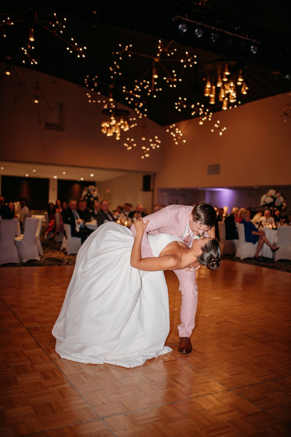 Couple in white dress and pink suit sharing dramatic dip dance move at wedding reception.