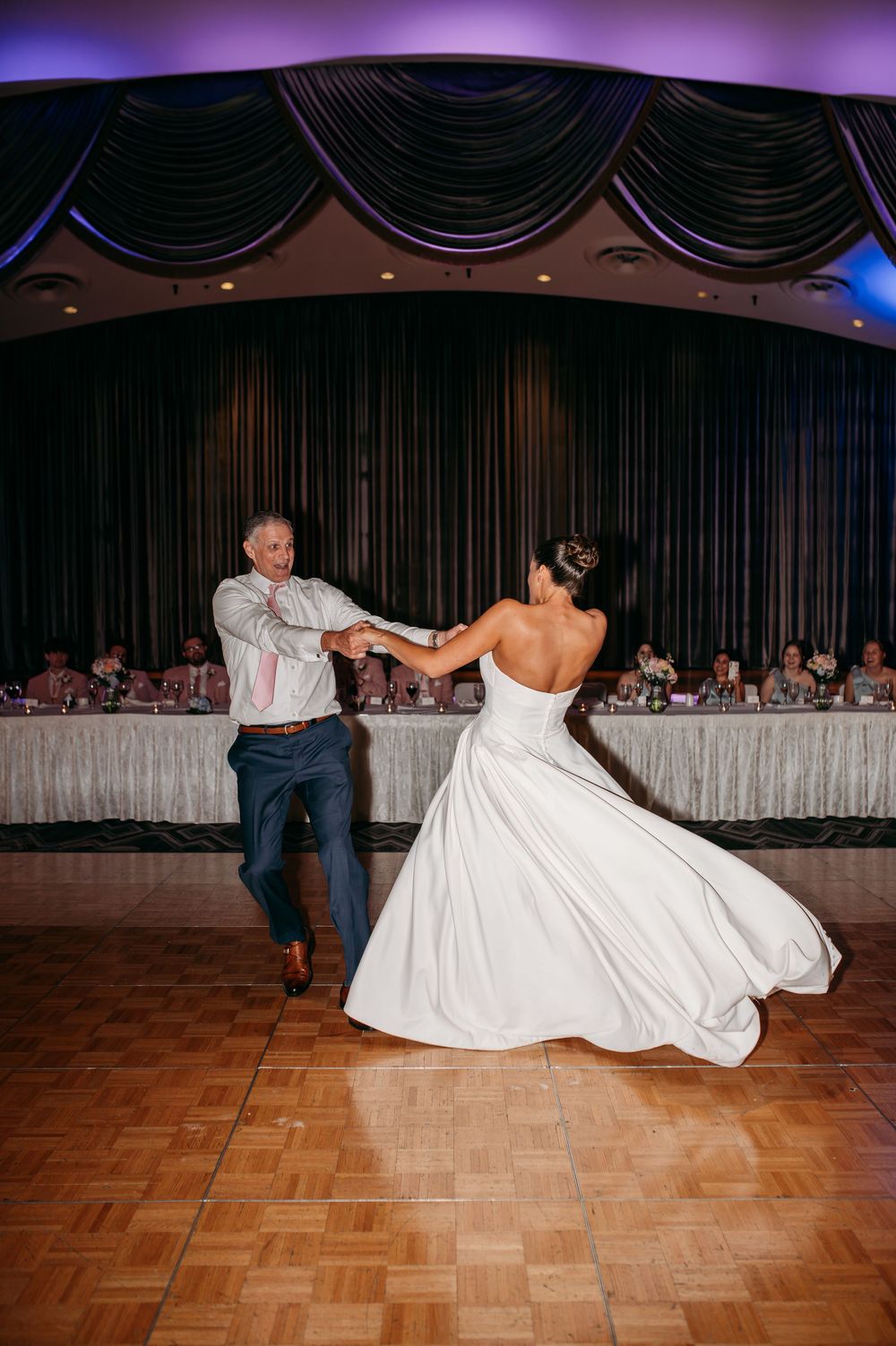 Couple dancing on a wooden floor during their wedding reception with elegant black curtains and decor in the background.