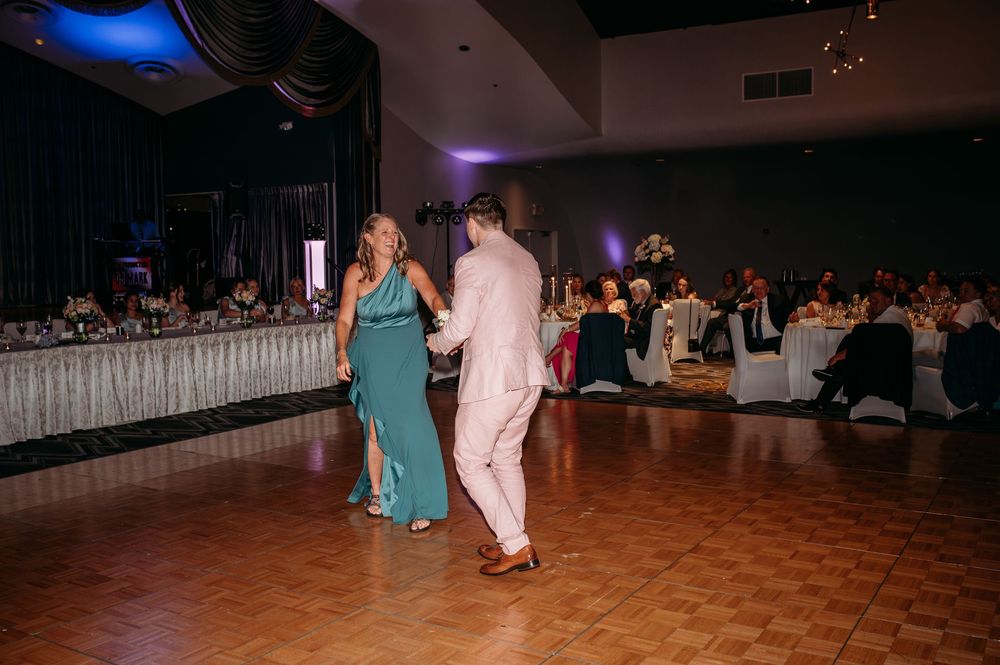 A couple dances together on a wooden floor during a formal event with decorated tables and guests in the background.