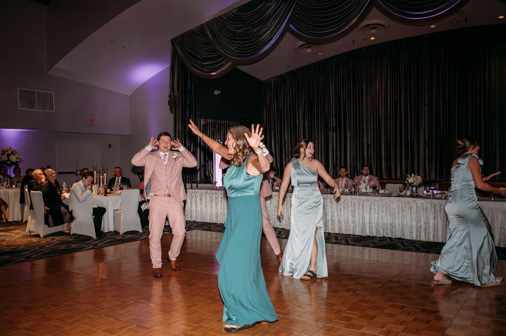 A group of wedding guests dance together on a dimly lit ballroom floor during a reception celebration.
