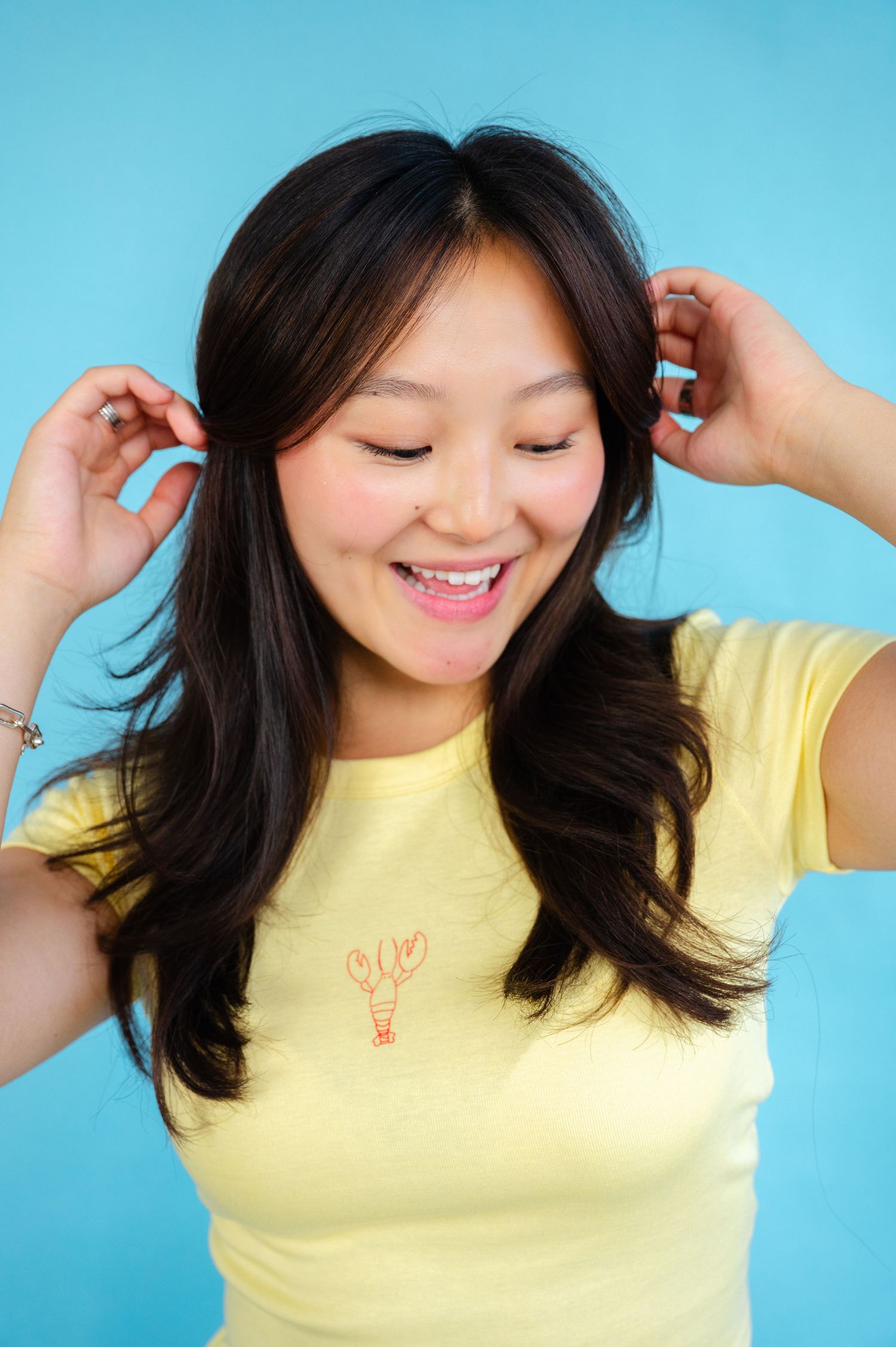 A person in a yellow t-shirt smiles joyfully against a blue background while playing with their long dark hair.