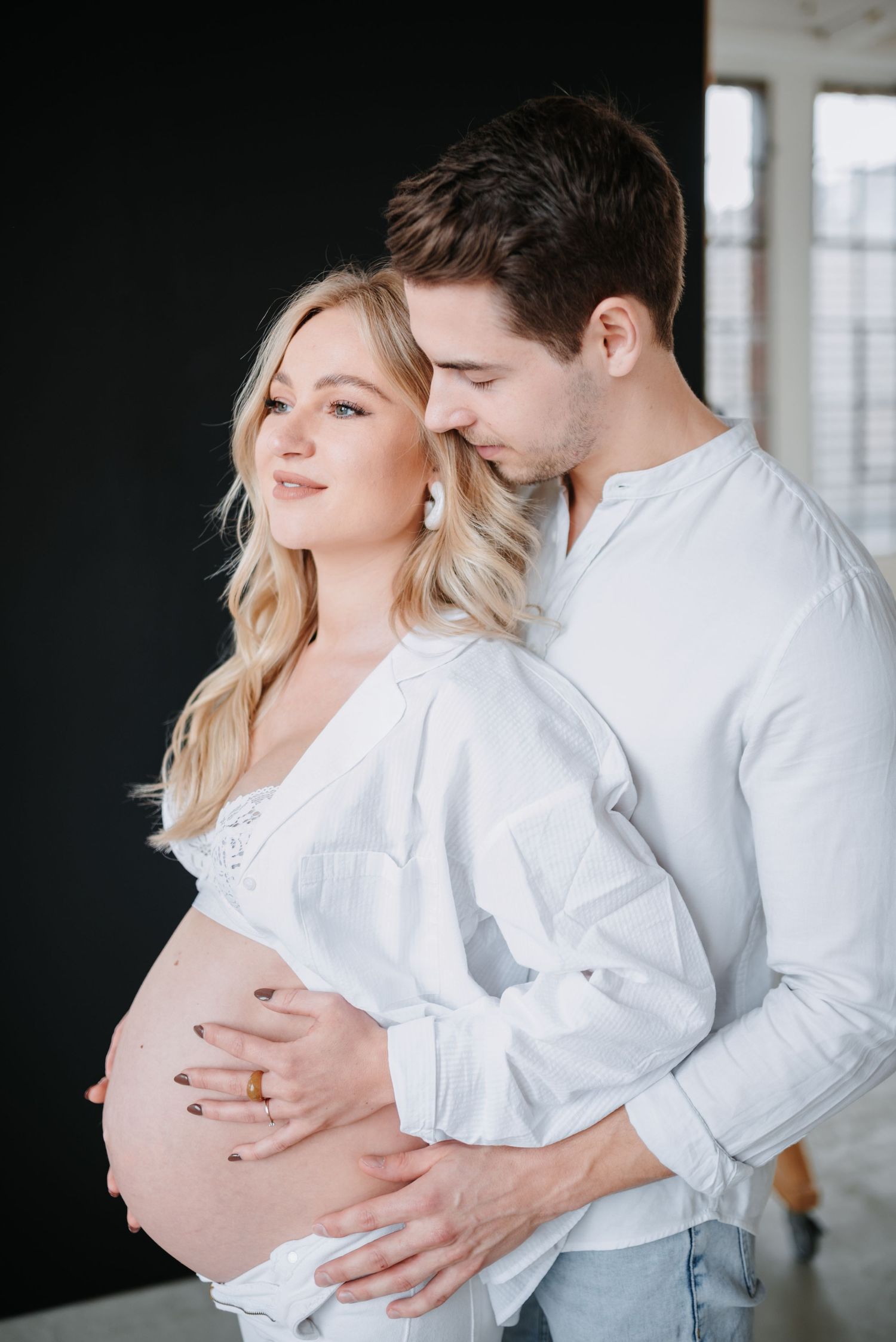 Intimate maternity photo of couple in white clothing embracing while showing pregnant belly in indoor setting.
