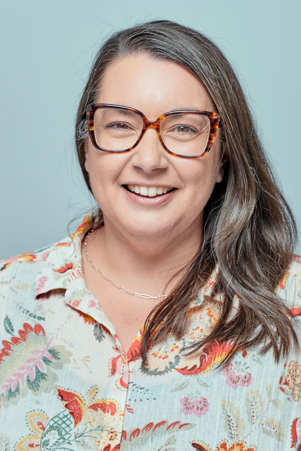 Professional portrait of someone wearing glasses and a floral print blouse against a light blue background.