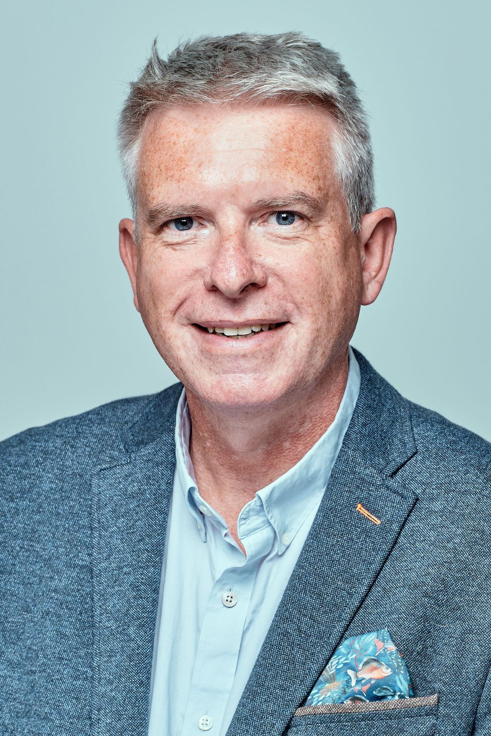 Professional headshot of a person in a blazer and light blue shirt against a blue backdrop.