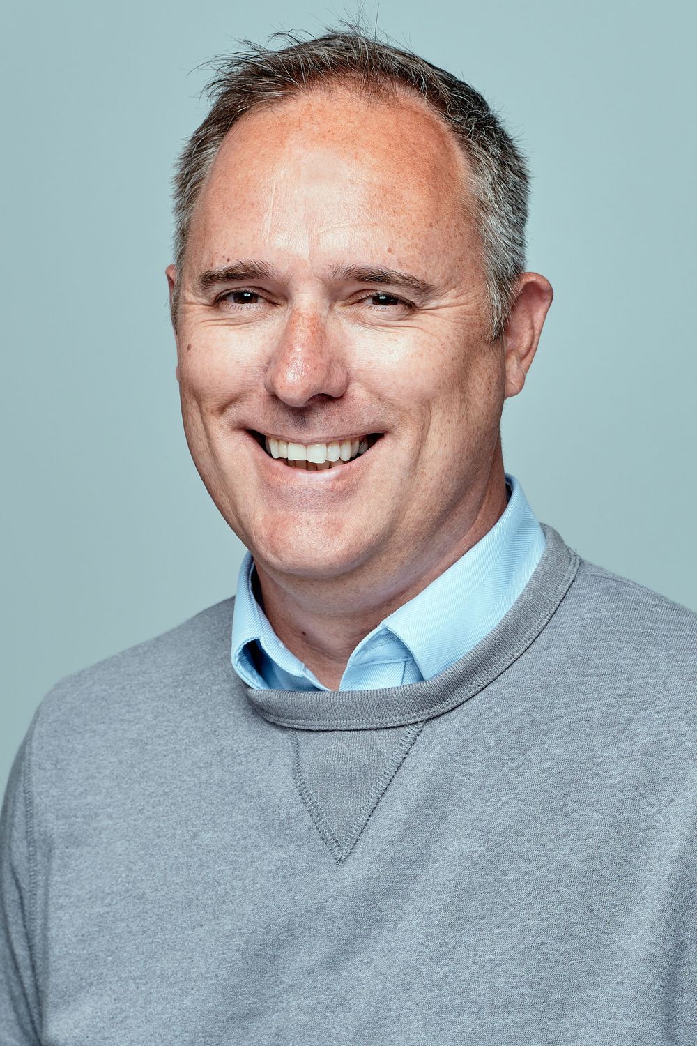 Progressive headshots of smiling person wearing light blue shirt and grey sweater against blue backdrop.