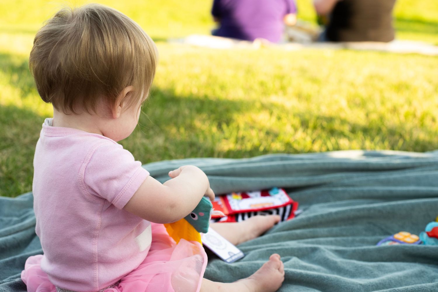 A baby in a pink sweater sits comfortably on a green blanket outdoors while playing with a toy.