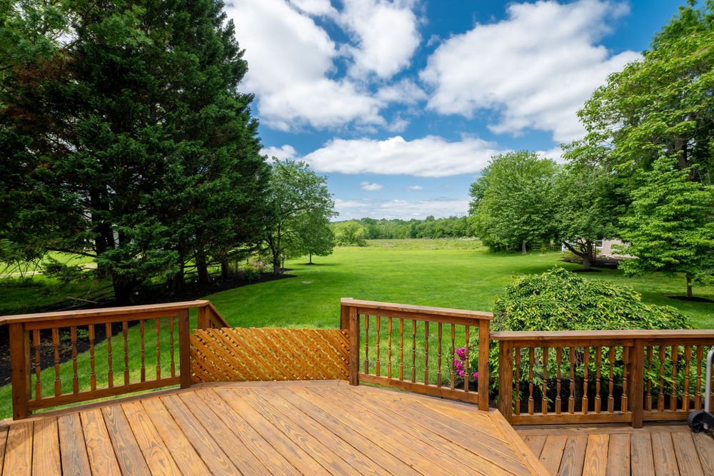 A wooden deck with railing overlooks a lush green lawn and tree-lined landscape under blue summer skies.