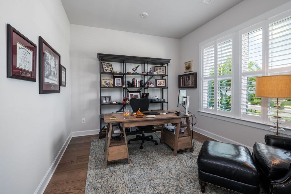 Modern home office featuring a dark wooden desk, bookshelf, leather chair and area rug in a bright room with plantation shutters.