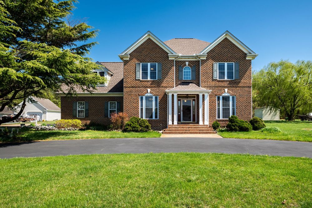 Elegant two-story brick colonial home with columns and dormers sits on a manicured lawn with curved driveway.