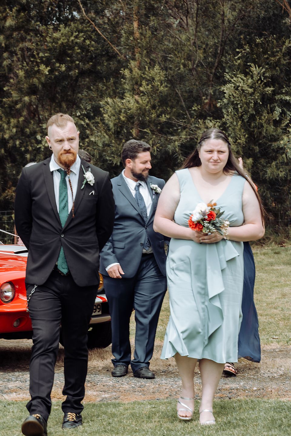 Wedding party poses playfully in a line on grass with black suits and green ties near outdoor foliage.