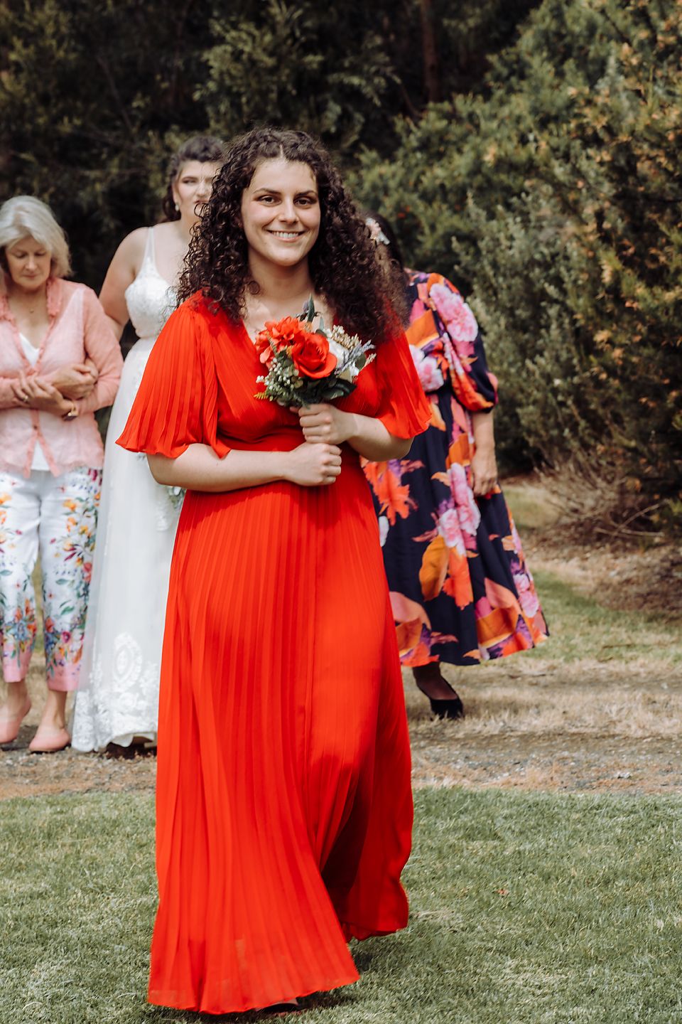 A wedding guest in a flowing red pleated dress holds a bouquet of red flowers at an outdoor garden ceremony.
