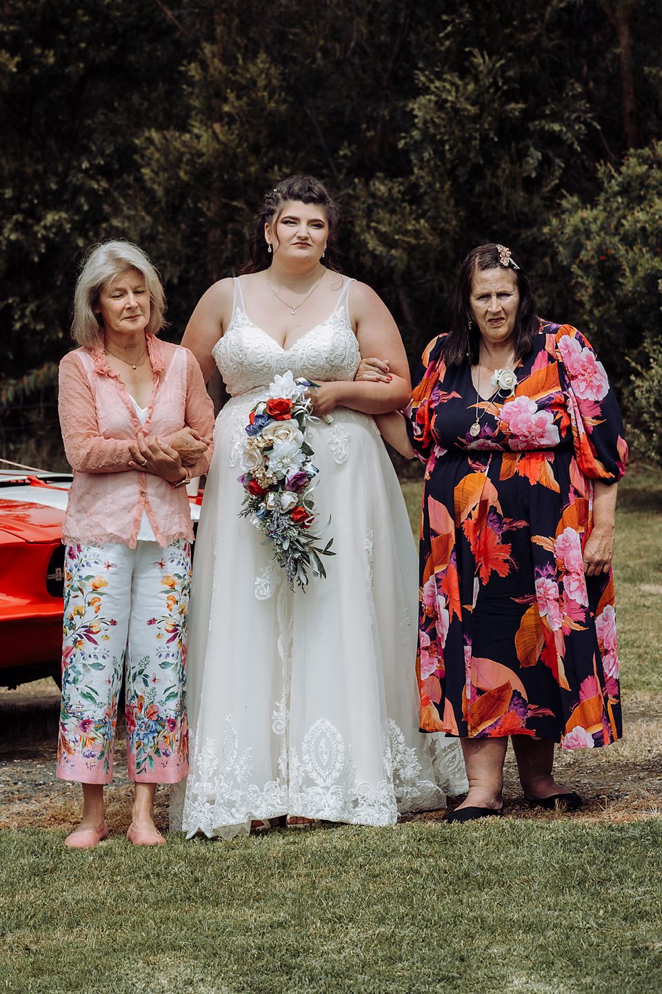 Wedding group poses sitting on grass featuring floral dresses and a white bridal gown with colorful bouquets.