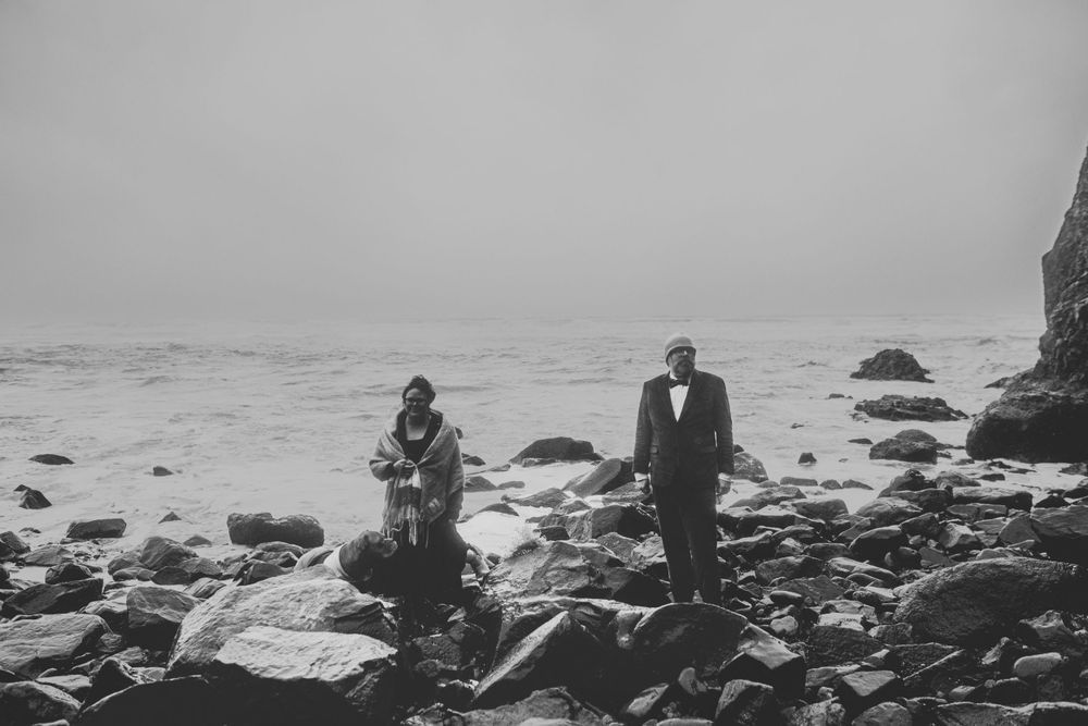 Two figures stand on rocky shoreline during moody overcast day by the ocean.