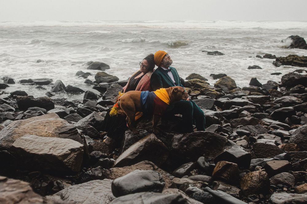 A couple wearing formal attire sits on rocky coastal terrain in moody overcast weather.